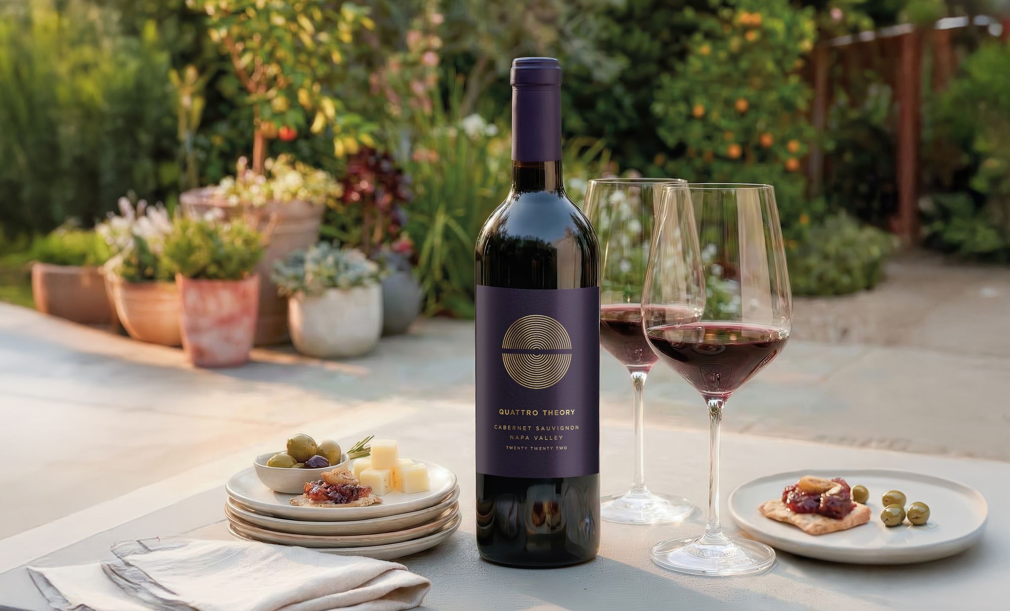 An outdoor patio table with a Quattro Theory Cabernet Sauvignon bottle, filled wine glasses, plates with food, napkins, and with potted plants and foliage in the background