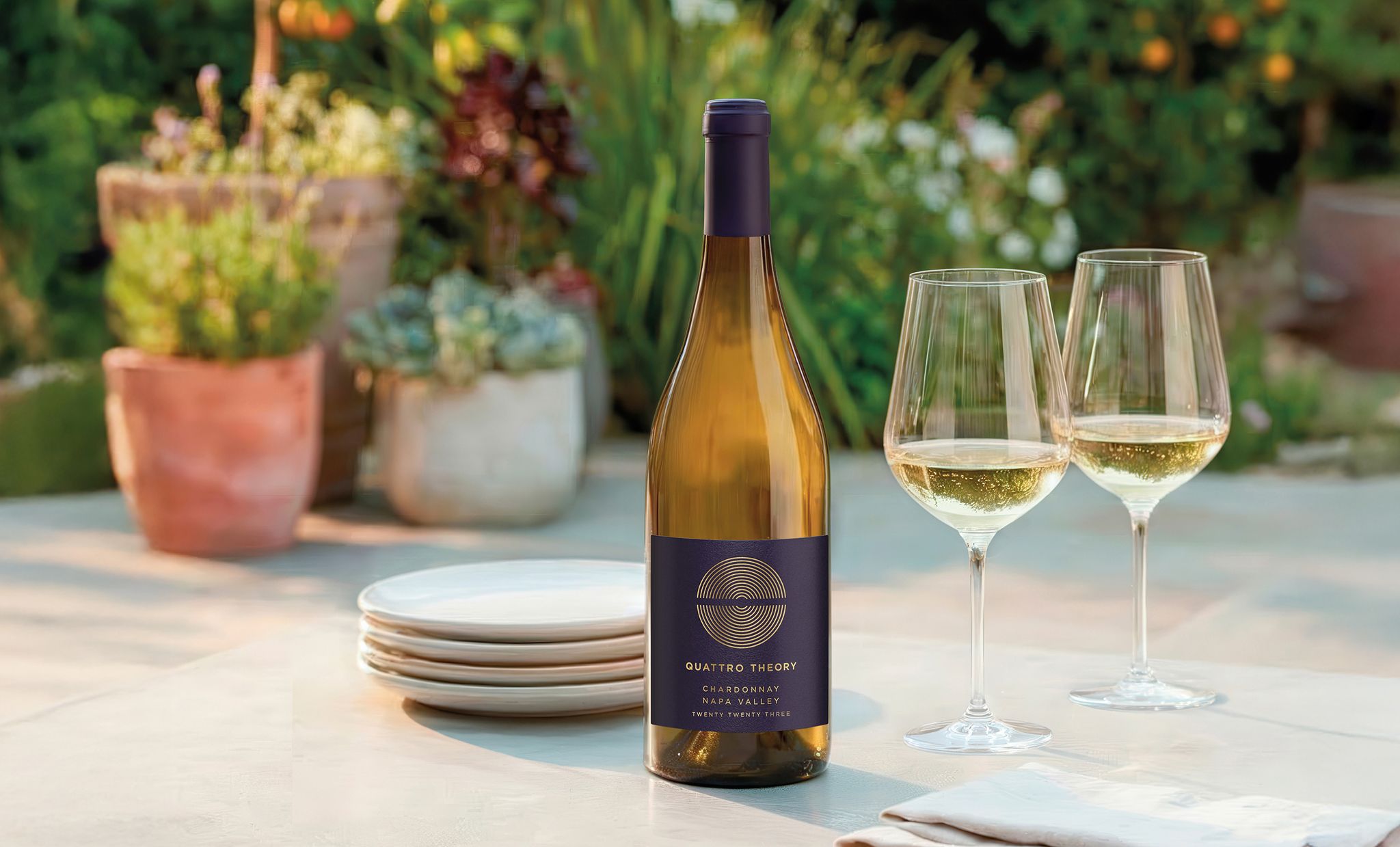 An outdoor patio table with a Quattro Theory Chardonnay wine bottle, filled wine glasses, plates and napkins, with potted plants and foliage in the background