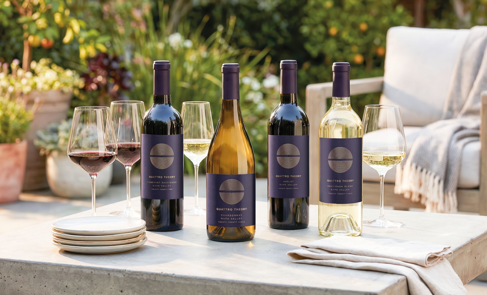 An outdoor patio table setting and chairs with Quattro Theory wine bottles, filled wine glasses, plates and napkins, with a chair and foliage in the background